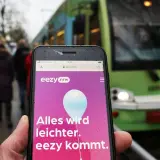 Ein Smartphone zeigt die "eezy.nrw" App mit der Botschaft "Alles wird leichter. eezy kommt." vor einer grünen Straßenbahn.