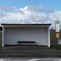 Ein Bushäuschen mit Bank und Haltestellenschild. Dieses Verkehrsmittel mit H-Symbol wartet auf Fahrgäste.