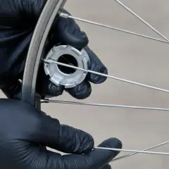 Hände mit schwarzen Handschuhen spannen die Speichen eines Fahrradrads mit einem Werkzeug.