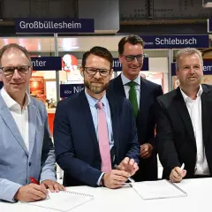 Vier Männer unterschreiben Verträge vor Schildern mit Ortsnamen wie Großbüllesheim. Die MOF Bedeutung wird hier wohl im geschäftlichen Kontext erklärt.