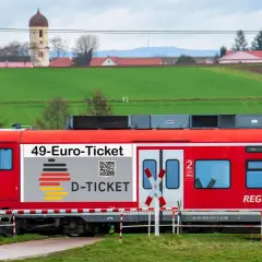 Rote Bahn mit "49-Euro-Ticket" Aufdruck fährt durch ländliche Gegend. Das rrx deutschlandticket macht Reisen günstig.