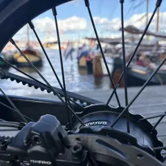 Nahaufnahme eines Fahrradrades mit Riemenantrieb und Nabenschaltung. Im Hintergrund liegen Boote im Hafen.