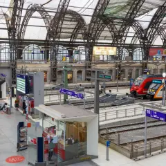 Menschen eilen durch eine Bahnhofshalle mit Zügen auf den Gleisen. Eine Tafel zeigt "Rauchfreier Bahnhof" und die Gleisnummern, Teil der Sackbahnhof Liste.