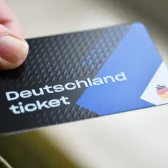 Ein Daumen hält ein Deutschlandticket. Das Ticket hat ein schwarzes, strukturiertes Design mit einem blauen und weißen Bereich.