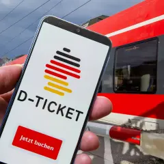 Smartphone zeigt D-Ticket App mit "Jetzt buchen" Button. Im Hintergrund rote Bahn in NRW.