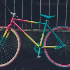 Ein bunt lackiertes Fahrrad mit pinken, gelben und türkisen Akzenten, das an einem Gitter lehnt.