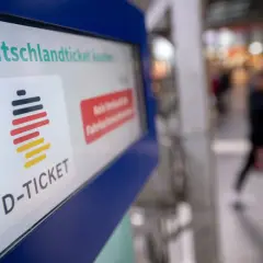 Das Deutschlandticket-Logo auf einem Bildschirm. Eine Person geht unscharf im Hintergrund vorbei. Vielleicht kann man das Deutschlandticket pausieren.