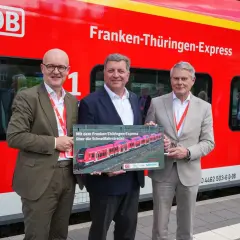 Drei Männer stehen vor einem roten Zug mit der Aufschrift "Franken-Thüringen-Express". Sie halten ein Schild mit einem Bild des Zuges.