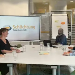Besprechung im Büro der Schlichtungsstelle Bahn. Mehrere Personen sitzen an einem Tisch, auf dem Bildschirm ist das Logo "Schlichtung Reise & Verkehr" zu sehen.