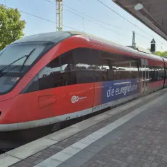 Roter Zug der euregiobahn br 643 steht am Bahnsteig. Grüne Bäume und blauer Himmel im Hintergrund.