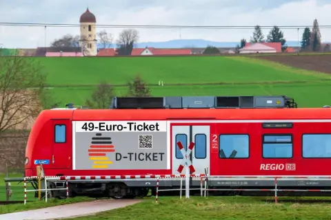 Rote Bahn mit "49-Euro-Ticket" Aufdruck fährt durch ländliche Gegend. Das rrx deutschlandticket macht Reisen günstig.