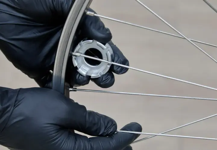 Hände mit schwarzen Handschuhen spannen die Speichen eines Fahrradrads mit einem Werkzeug.