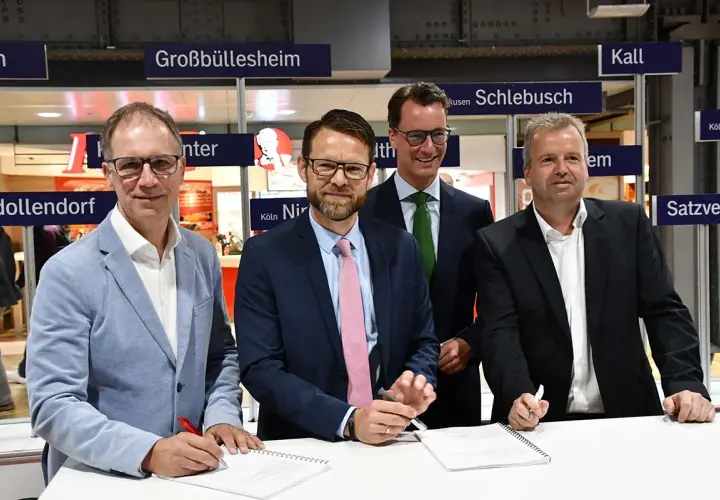 Vier Männer unterschreiben Verträge vor Schildern mit Ortsnamen wie Großbüllesheim. Die MOF Bedeutung wird hier wohl im geschäftlichen Kontext erklärt.