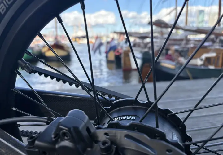 Nahaufnahme eines Fahrradrades mit Riemenantrieb und Nabenschaltung. Im Hintergrund liegen Boote im Hafen.
