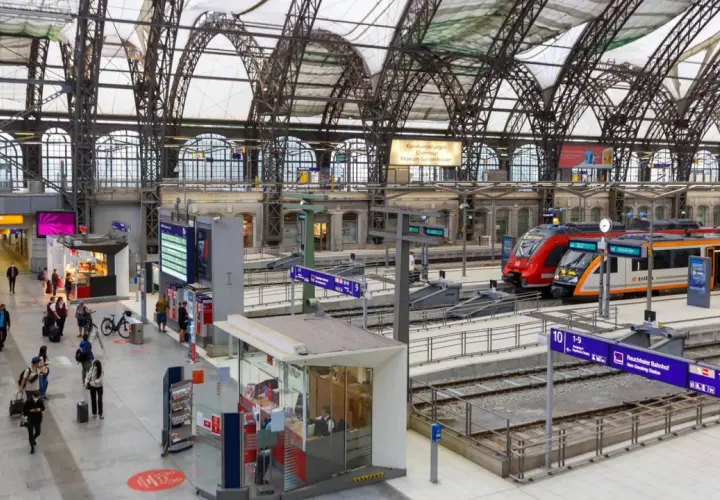 Menschen eilen durch eine Bahnhofshalle mit Zügen auf den Gleisen. Eine Tafel zeigt "Rauchfreier Bahnhof" und die Gleisnummern, Teil der Sackbahnhof Liste.