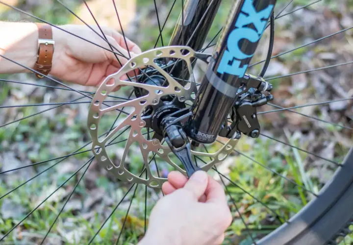 Ein Mann mit braunem Lederarmband schließt die **steckachse vs schnellspanner** am Fahrrad. Die Fox-Gabel und die Shimano-Bremse sind sichtbar.