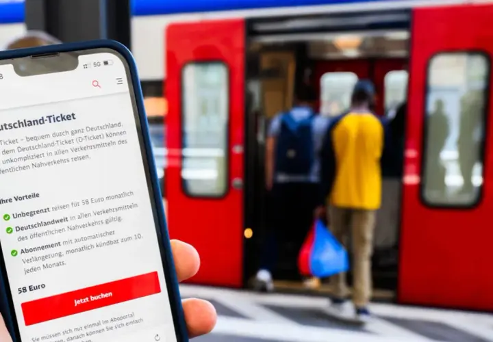 Smartphone zeigt Deutschland-Ticket mit Vorteilen: unbegrenzt reisen, deutschlandweit gültig, 58 Euro/Monat. Bahnsteig mit einsteigenden Reisenden im Hintergrund.