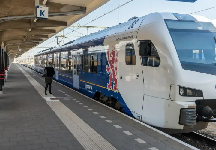 Eine Frau wartet auf dem Bahnsteig auf den Zug. Der blaue Arriva-Zug mit dem roten Löwenwappen ist ein Teil des Nahverkehrs in den Niederlanden.