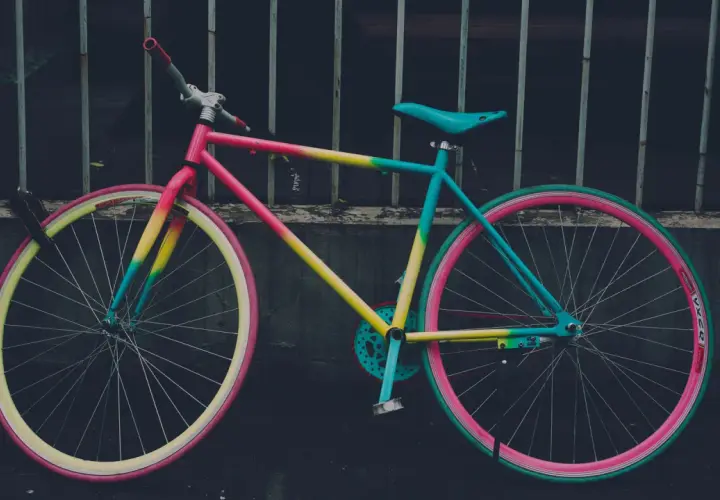Ein bunt lackiertes Fahrrad mit pinken, gelben und türkisen Akzenten, das an einem Gitter lehnt.