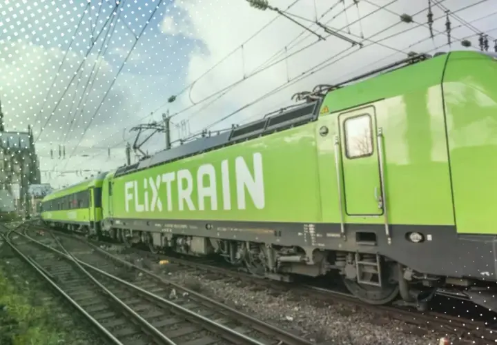 Ein grüner FlixTrain fährt auf Gleisen, im Hintergrund ist eine Kathedrale zu sehen. Perfekt für eine Reise mit dem Deutschlandticket.