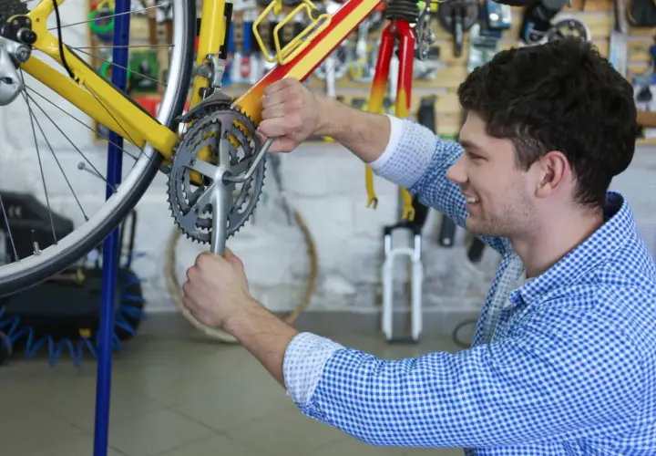 Ein Mann restauriert ein gelbes Fahrrad in einer Werkstatt. Er schraubt mit einem Werkzeug an der Kurbel.