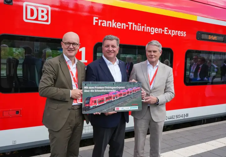 Drei Männer stehen vor einem roten Zug mit der Aufschrift "Franken-Thüringen-Express". Sie halten ein Schild mit einem Bild des Zuges.