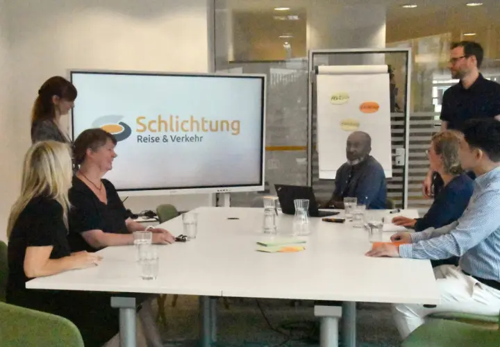Besprechung im Büro der Schlichtungsstelle Bahn. Mehrere Personen sitzen an einem Tisch, auf dem Bildschirm ist das Logo "Schlichtung Reise & Verkehr" zu sehen.