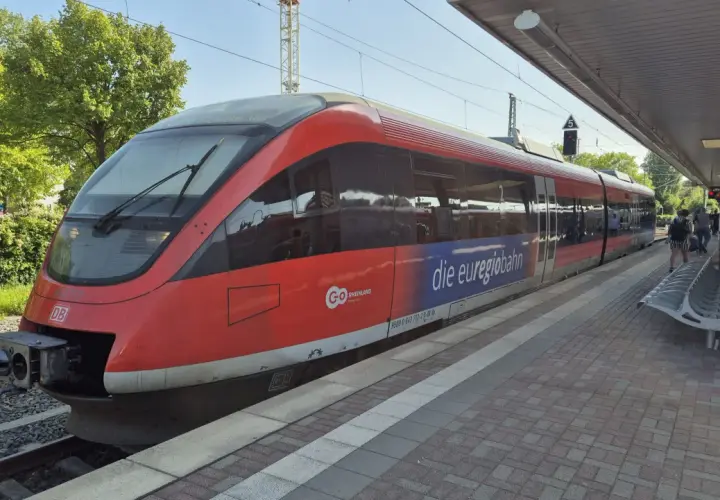 Roter Zug der euregiobahn br 643 steht am Bahnsteig. Grüne Bäume und blauer Himmel im Hintergrund.
