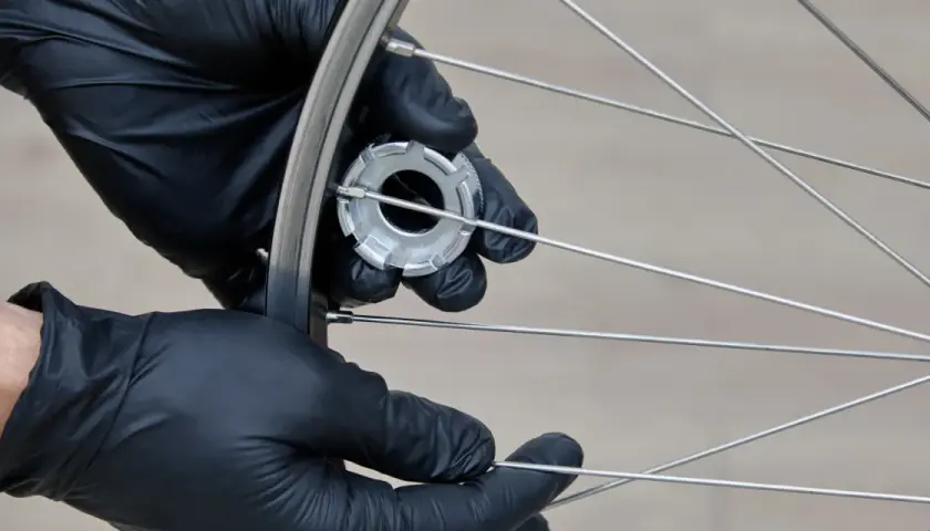 Hände mit schwarzen Handschuhen spannen die Speichen eines Fahrradrads mit einem Werkzeug.
