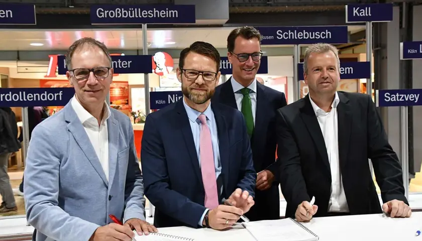Vier Männer unterschreiben Verträge vor Schildern mit Ortsnamen wie Großbüllesheim. Die MOF Bedeutung wird hier wohl im geschäftlichen Kontext erklärt.