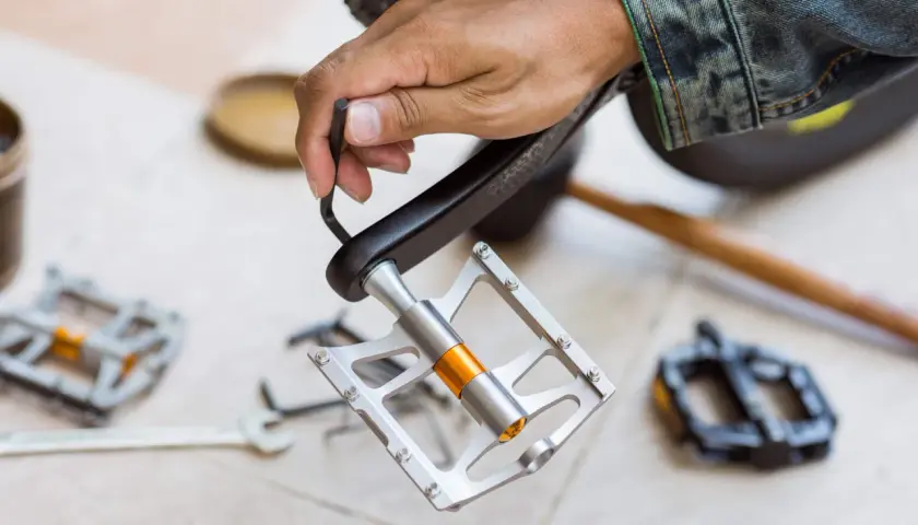 Hand mit Inbusschlüssel montiert neue Fahrradpedale. Werkzeug und alte Pedale liegen bereit.