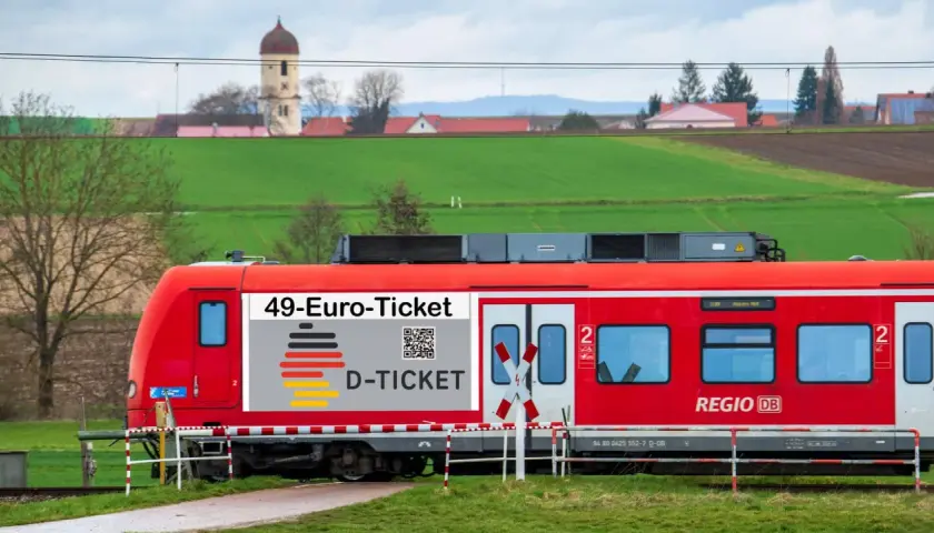 Rote Bahn mit "49-Euro-Ticket" Aufdruck fährt durch ländliche Gegend. Das rrx deutschlandticket macht Reisen günstig.
