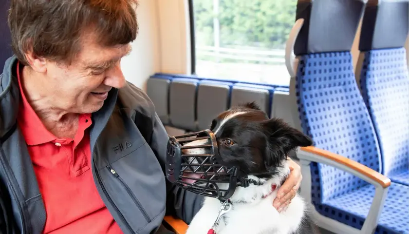 Ein Mann mit seinem Hund im Zug in NRW. Der Hund trägt einen Maulkorb und schaut freundlich.