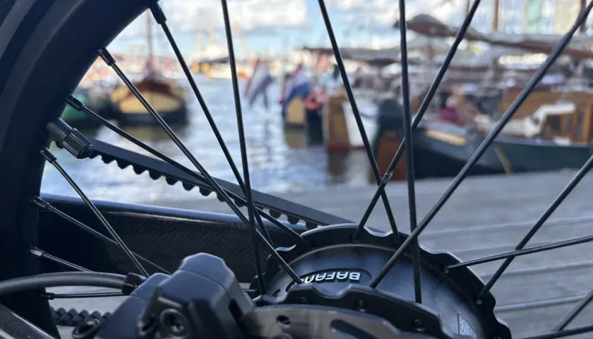 Nahaufnahme eines Fahrradrades mit Riemenantrieb und Nabenschaltung. Im Hintergrund liegen Boote im Hafen.