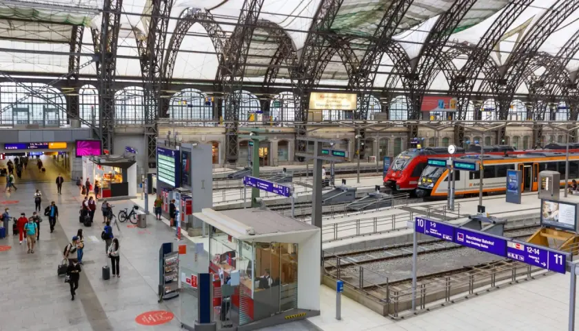 Menschen eilen durch eine Bahnhofshalle mit Zügen auf den Gleisen. Eine Tafel zeigt "Rauchfreier Bahnhof" und die Gleisnummern, Teil der Sackbahnhof Liste.