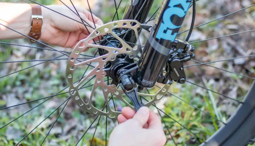 Ein Mann mit braunem Lederarmband schließt die **steckachse vs schnellspanner** am Fahrrad. Die Fox-Gabel und die Shimano-Bremse sind sichtbar.