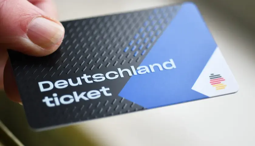 Ein Daumen hält ein Deutschlandticket. Das Ticket hat ein schwarzes, strukturiertes Design mit einem blauen und weißen Bereich.