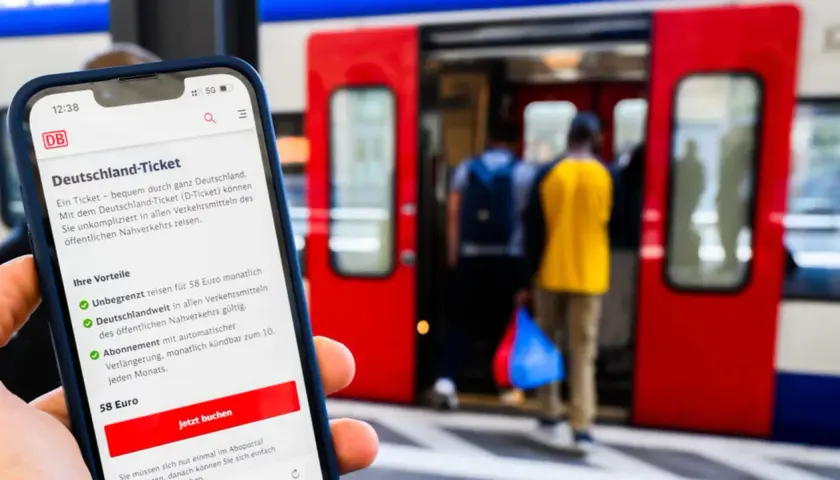 Smartphone zeigt Deutschland-Ticket mit Vorteilen: unbegrenzt reisen, deutschlandweit gültig, 58 Euro/Monat. Bahnsteig mit einsteigenden Reisenden im Hintergrund.