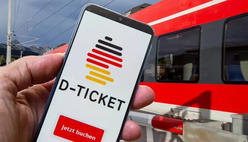 Smartphone zeigt D-Ticket App mit "Jetzt buchen" Button. Im Hintergrund rote Bahn in NRW.
