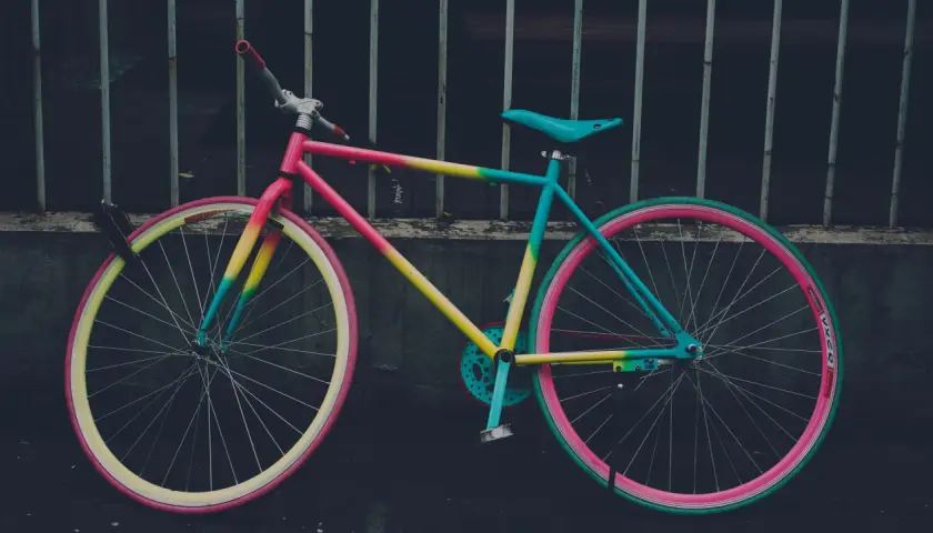 Ein bunt lackiertes Fahrrad mit pinken, gelben und türkisen Akzenten, das an einem Gitter lehnt.