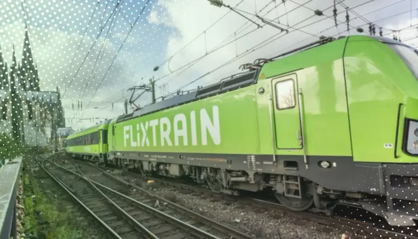 Ein grüner FlixTrain fährt auf Gleisen, im Hintergrund ist eine Kathedrale zu sehen. Perfekt für eine Reise mit dem Deutschlandticket.