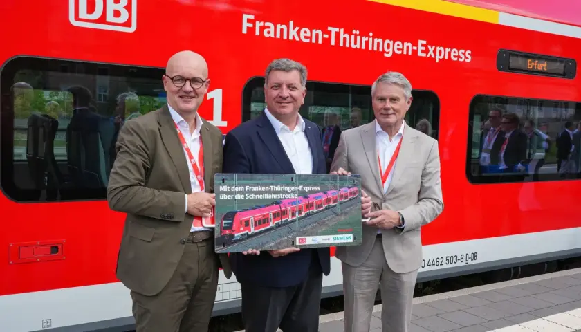 Drei Männer stehen vor einem roten Zug mit der Aufschrift "Franken-Thüringen-Express". Sie halten ein Schild mit einem Bild des Zuges.