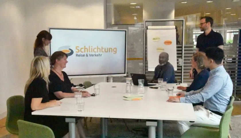 Besprechung im Büro der Schlichtungsstelle Bahn. Mehrere Personen sitzen an einem Tisch, auf dem Bildschirm ist das Logo "Schlichtung Reise & Verkehr" zu sehen.