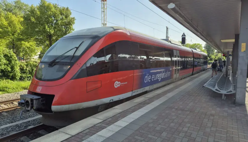Roter Zug der euregiobahn br 643 steht am Bahnsteig. Grüne Bäume und blauer Himmel im Hintergrund.