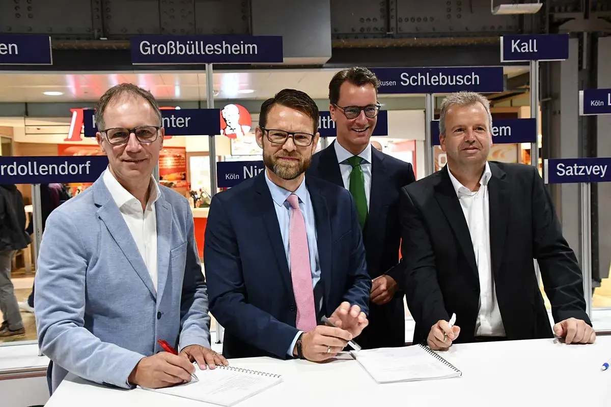 Vier Männer unterschreiben Verträge vor Schildern mit Ortsnamen wie Großbüllesheim. Die MOF Bedeutung wird hier wohl im geschäftlichen Kontext erklärt.