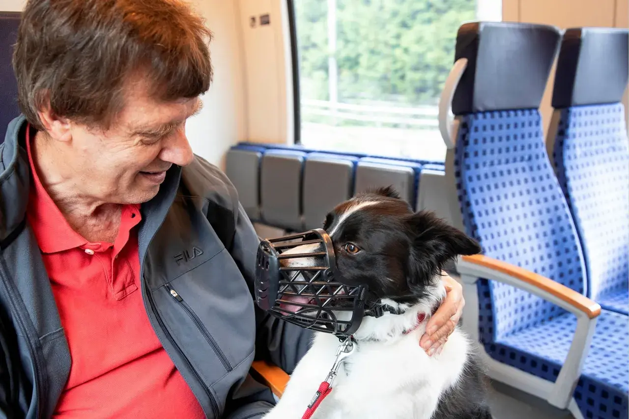 Ein Mann mit seinem Hund im Zug in NRW. Der Hund trägt einen Maulkorb und schaut freundlich.