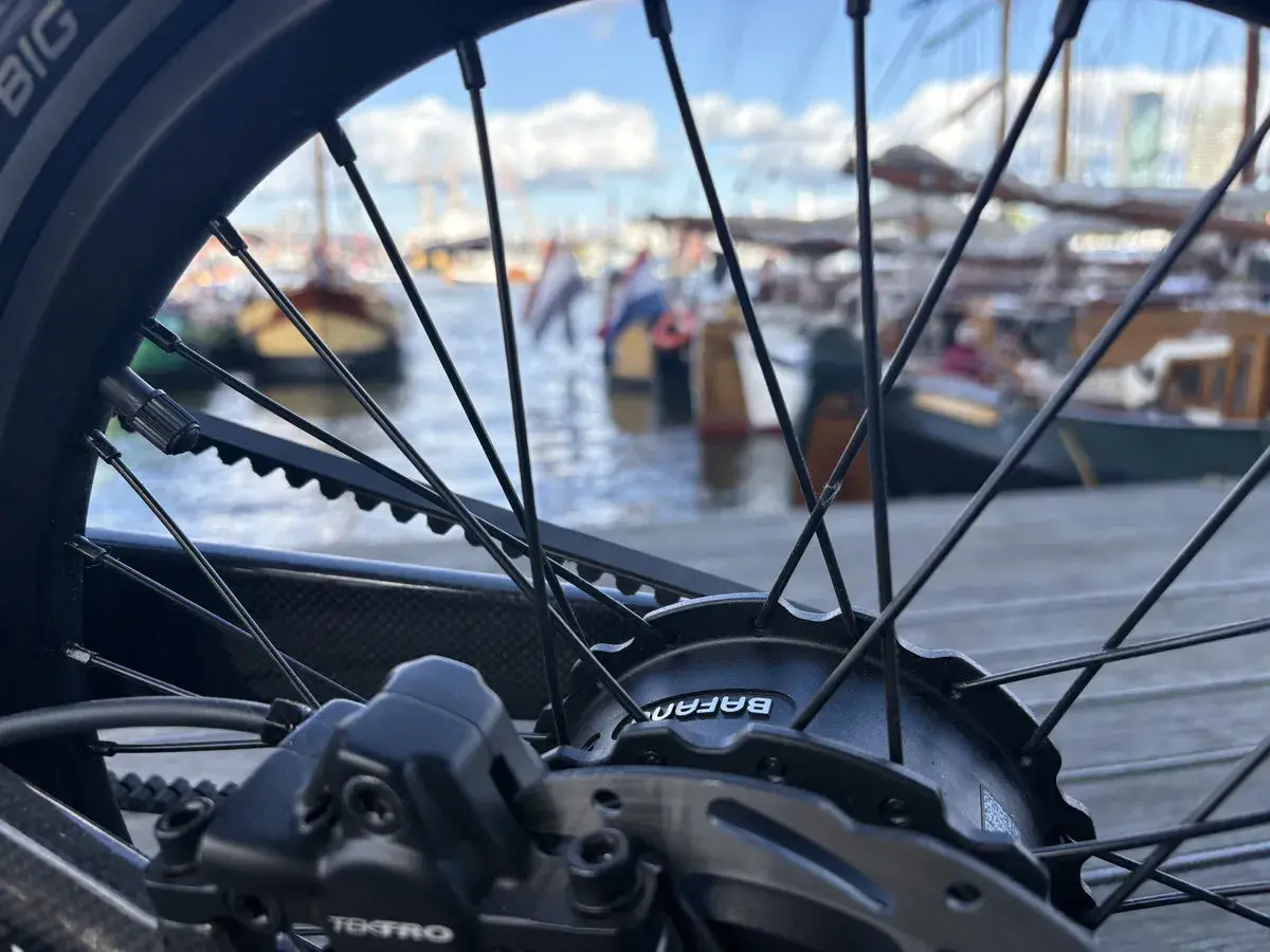 Nahaufnahme eines Fahrradrades mit Riemenantrieb und Nabenschaltung. Im Hintergrund liegen Boote im Hafen.