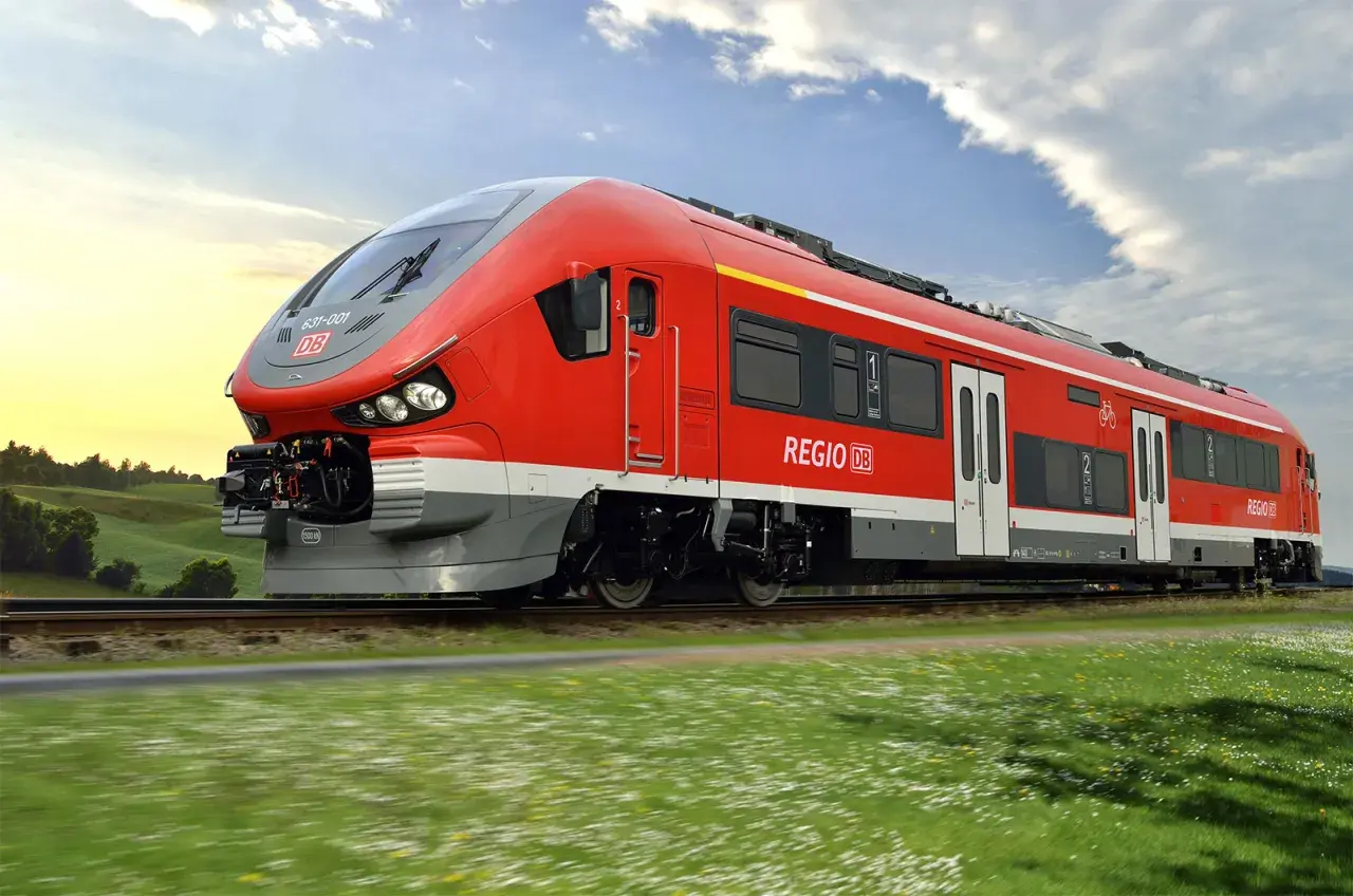Roter Pesa-Zug der Deutschen Bahn fährt durch eine grüne Landschaft unter blauem Himmel.