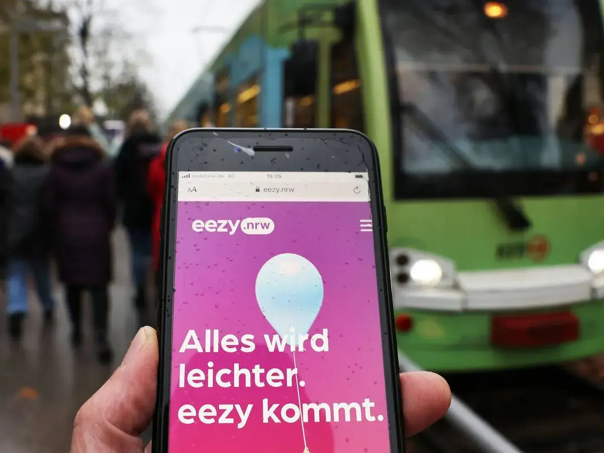 Ein Smartphone zeigt die "eezy.nrw" App mit der Botschaft "Alles wird leichter. eezy kommt." vor einer grünen Straßenbahn.