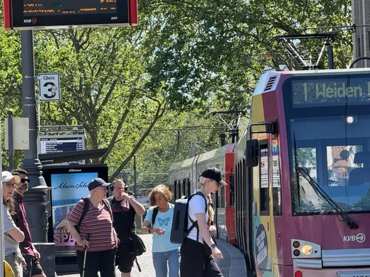 Menschen warten an Gleis 3 auf die Bahn. Die Anzeige zeigt "1 Weiden", was die einfache Fahrt Bedeutung für die Reisenden verdeutlicht.
